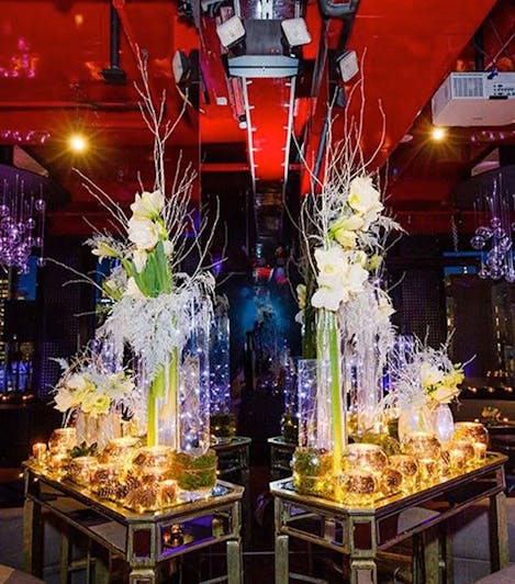 A pair of tall glass vases, stuffed with white floral arrangements, are positioned atop matching glass tabletops